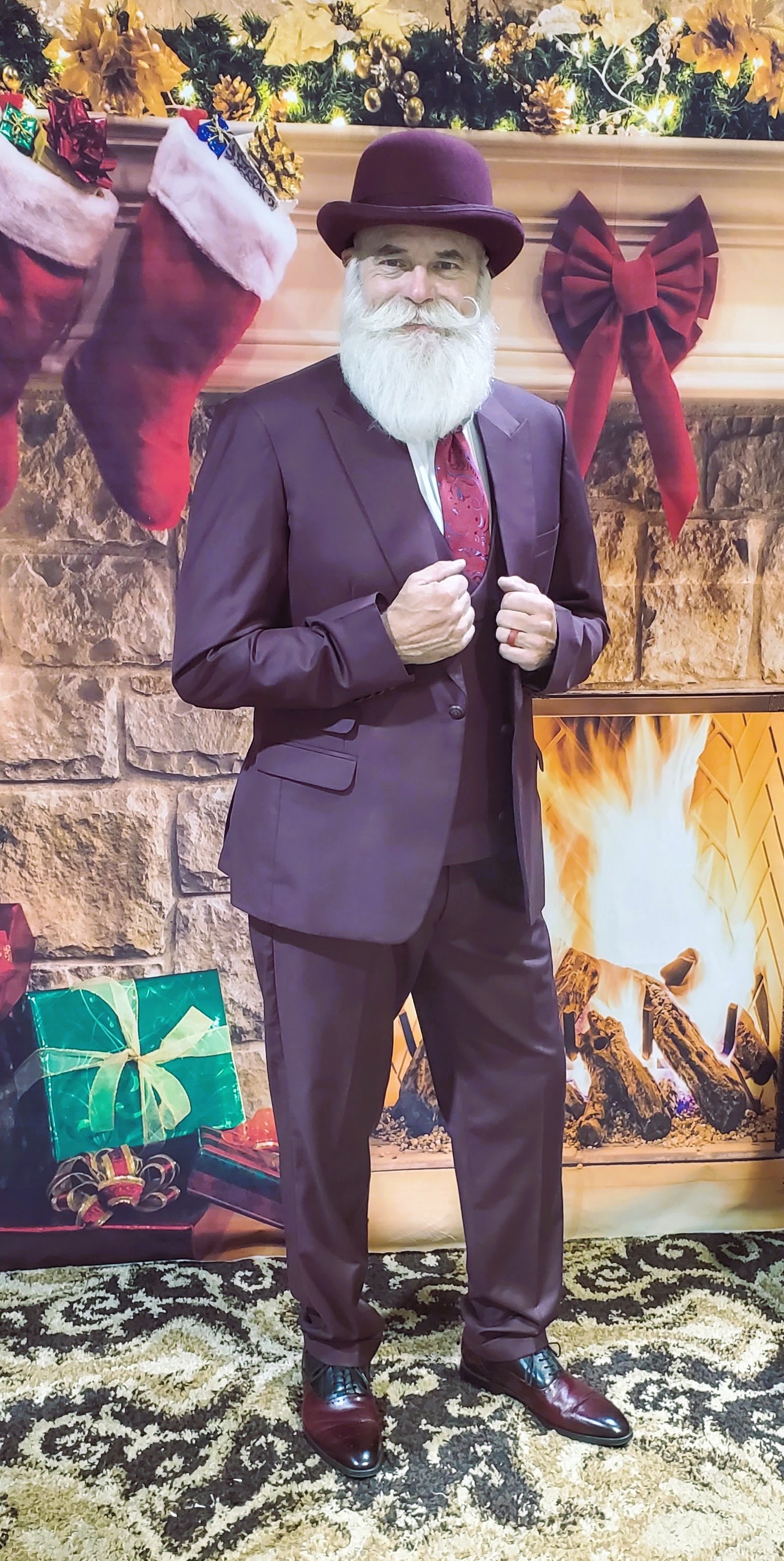 Santa in a wine-colored suit and hat before a Christmas fireplace