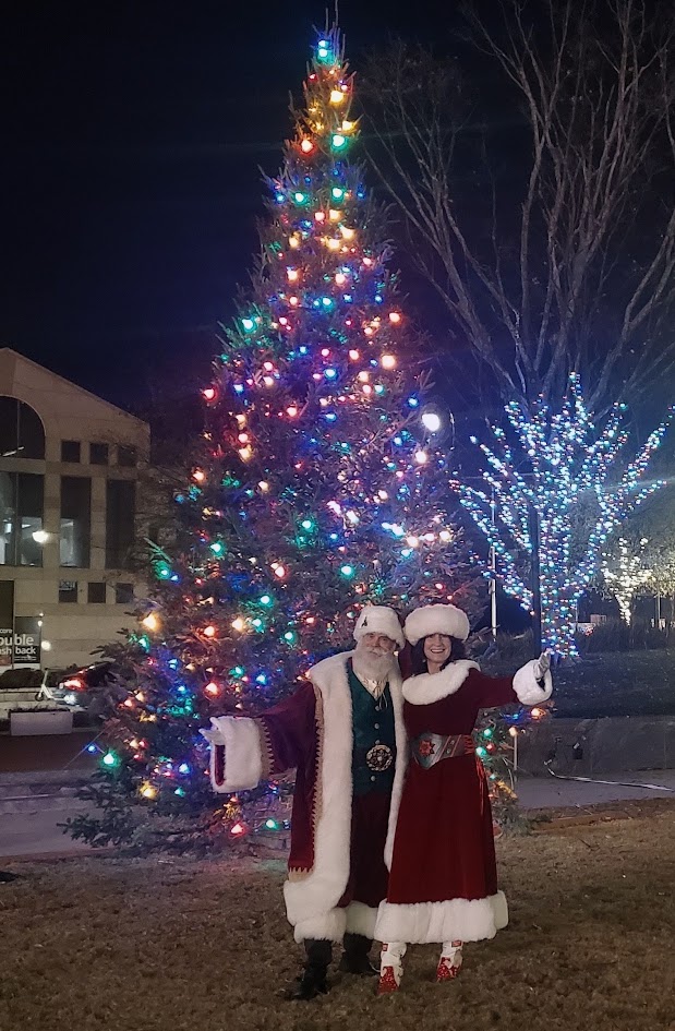 Santa and Mrs. Claus before a brightly lit community Christmas tree